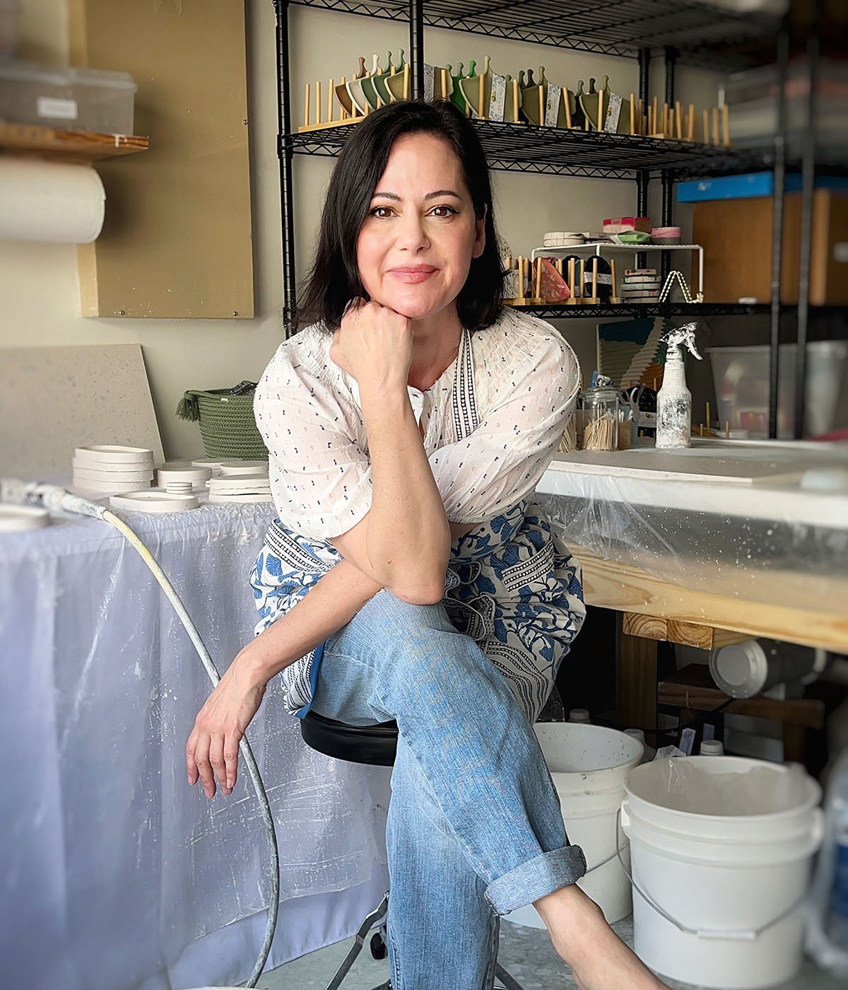 Photo of the artist in her studio with blue jeans and a summer top on. She has a French bob haircut, is barefoot with her head resting on her chin. Her housewares materials are around her as she looks like she is working on her terrazzo housewares..